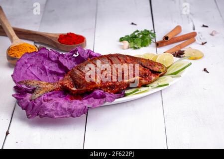 Pomfret braten in einem weißen Teller angeordnet garniert mit lila Kohlblatt und Gurkenscheiben auf weißem strukturierten Hintergrund. Stockfoto