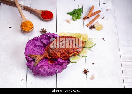 Pomfret braten in einem weißen Teller angeordnet garniert mit lila Kohlblatt und Gurkenscheiben auf weißem strukturierten Hintergrund. Stockfoto