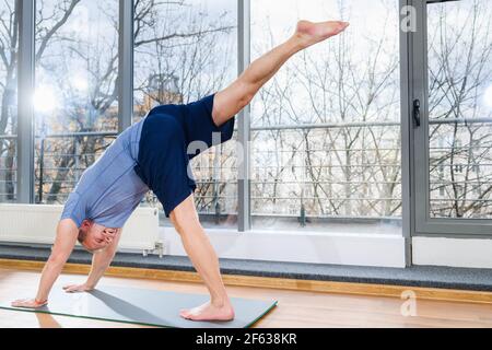 Mittelalter positiver sportlicher Mann trainieren indoor im hellen sonnigen Fitnessstudio mit Panoramafenster, Stretch-Beine, Rückansicht. Nach Unten Hund Pose Stockfoto