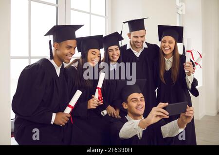 Gruppe von glücklichen internationalen Studenten mit Diplomen unter Selfie mit Smartphone im Klassenzimmer. Stockfoto