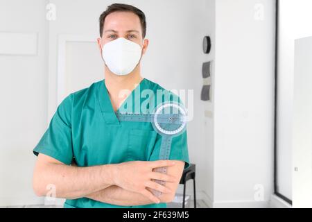 Freundliche männliche Physiotherapeut posiert mit Werkzeugen in der modernen Klinik trägt Schutzmaske Gesicht während Coronavirus Pandemie. Hochwertige Fotos Stockfoto