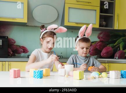 Ein kleiner Junge und ein Mädchen mit Kaninchenohren auf dem Kopf malen Ostereier, die zu Hause in der Küche sitzen Stockfoto