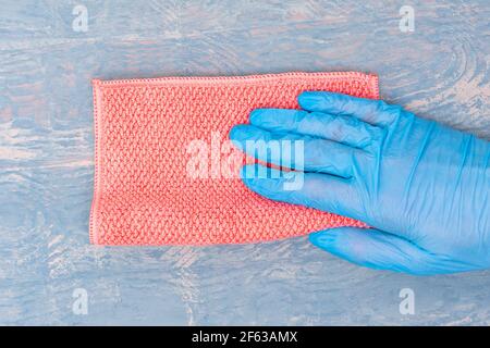 Hand in einem blauen Gummihandschuh wischt den Staub mit einem rosa Lappen auf einem blauen Holzhintergrund, kopieren Raum, Nahaufnahme. Haus, Haushaltskonzept. Stockfoto