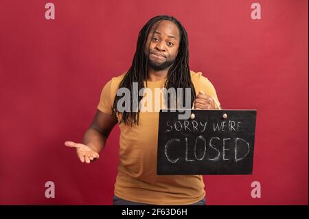 Enttäuscht junge afroamerikanische Kerl mit Dreadlocks stehen isoliert auf der roten Wand, hält schwarze Platte mit geschrieben wir sind geschlossen Text, unglücklich, die Nachrichten zu informieren, Geschäft kämpfen in Pandemie Stockfoto