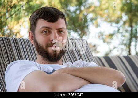 Nahaufnahme Porträt von jungen Erwachsenen cuacasian bärtigen Mann genießen Chillen auf dem Sofa im Hof im Freien an sonnigen warmen Sommertag. Glücklich lächelnde männliche Person Stockfoto