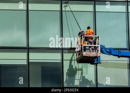 Fensterputzer, die hohe Bürofenster mit einer Kirschenpflücker-Plattform waschen. Arbeiter, die eine Luftarbeitsbühne benutzen, um auf hohe Gebäude zu gelangen. Wasserversorgte Stange Stockfoto