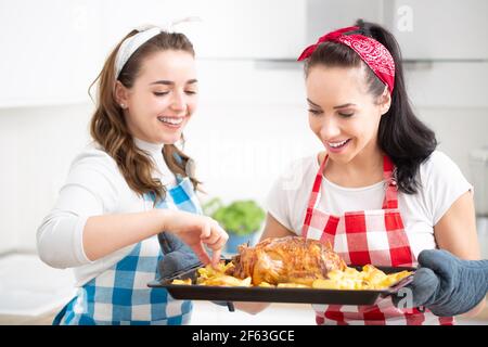 Zwei junge Hausfrauen in karierten Schürzen überprüfen und probieren frisch gebratenes Huhn und Kartoffeln in der Küche. Stockfoto