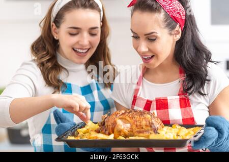 Zwei junge Hausfrauen in karierten Schürzen überprüfen und probieren frisch gebratenes Huhn und Kartoffeln in der Küche. Stockfoto
