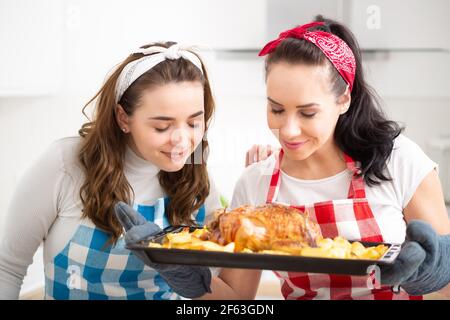 Zwei junge Frauen riechen frisch gebackenes Huhn mit Kartoffeln auf einem Backblech Stockfoto