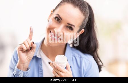 Frau hält Gesichtscreme aus einer weißen Verpackung an ihrem Finger genommen, lächelnd, Creme auch auf ihr Gesicht aufgetragen. Stockfoto