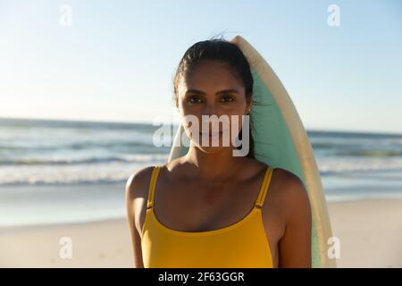 Porträt einer Mixed Race Frau mit Surfbrett am Strand Blick auf die Kamera Stockfoto