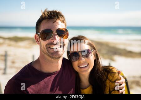 Porträt eines glücklichen kaukasischen Paares, das am Strand bei der sitzt Meer umarmt trägt Sonnenbrille lächelnd Stockfoto