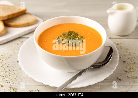 Hausgemachte Kürbis- und Karottensuppe in einer weißen Schüssel mit Sahne und Samen schließen sich auf weißem Holz rustikalen Hintergrund. Stockfoto