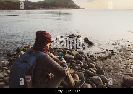 Ein junger Mann in warmen Kleidern sitzt auf dem Stein, trinken heißen Tee oder Kaffee aus wiederverwendbaren Thermoskanne und genießen Meerblick. Entspannen Sie sich bei einem Spaziergang auf Stockfoto