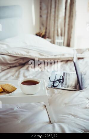 Weißes Holztablett mit einer Tasse heißen Tee oder Kaffee und Keksen mit einem Laptop auf dem Hintergrund. Frühstück von Geschäftsreisenden. Morgendliche Gewohnheiten von Stockfoto