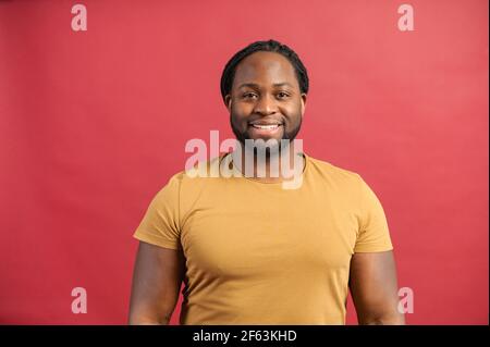 Freundlich und positiv afroamerikanisch mit Dreadlocks Kerl trägt helle T-Shirt schaut auf Kamera und lächelt isoliert auf rotem Hintergrund, glücklich dunkelhäutig Mann fühlt sich gut, hat Glückstag heute Stockfoto