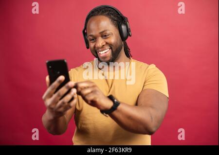 Urkomischer afroamerikanischer Mann, der Kopfhörer mit einem Smartphone trägt, seine Freizeit online verbringt, Videos anschaut, Musik hört und lächelt, glücklicher Schwarzer mit Ohrhörern hält das Telefon, isoliert auf Rot Stockfoto