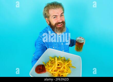 Lächelnder Mann mit Pommes Frites und Bier. Pommes Frites. Bärtiger Rüde mit Pommes Frites und Ketchup auf dem Teller. Stockfoto