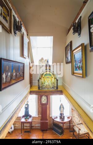 Blick auf die Treppe von der oberen Etage mit Gemälden an den Wänden und einem kleinen Zwischengeschoss, das mit einer Miniaturkirche in einem alten Haus aus dem Jahr 1790 dekoriert ist. Stockfoto