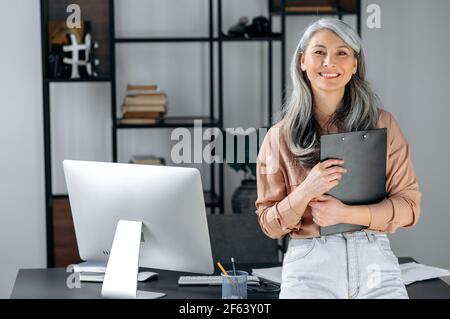 Porträt einer erfolgreichen, grauhaarigen asiatischen Geschäftsfrau mittleren Alters, die im modernen Büro steht, in stilvoller Kleidung gekleidet, Ordner in den Händen hält, die Kamera anschaut und freundlich lächelt Stockfoto