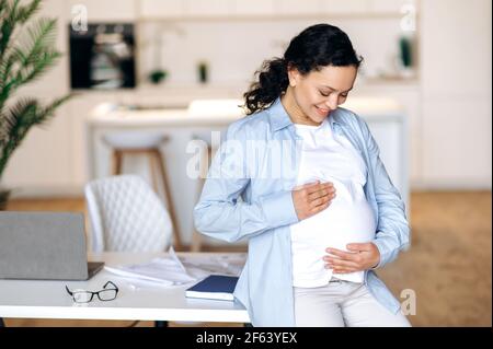 Schwanger. Nette, fröhliche, schwanger-reife Frau, gemischtes Rennen, Manager, Designer oder Agent, die aus der Ferne arbeiten, stilvolle Kleidung tragen, auf einem Tisch sitzen und den Bauch streicheln und glücklich lächeln Stockfoto