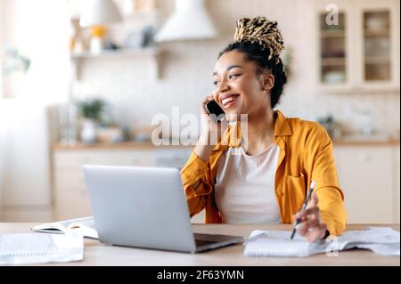 Glückliche erfolgreiche vielbeschäftigte junge afroamerikanische Frau, Maklerin, Managerin oder Freiberuflerin, die aus der Ferne arbeitet, unterhalten sich angenehm mit dem Kunden oder Mitarbeiter am Telefon und schauen lächelnd freundlich weg Stockfoto