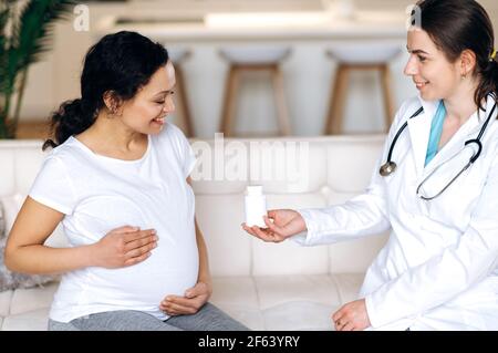 Gesundes Schwangerschaftskonzept. Ein Arzt verschreibt einer glücklichen, gemischten Schwangeren Medikamente oder Vitamine, die zum Arzttermin, zu Hause oder im Krankenhaus auf einem Sofa sitzen Stockfoto