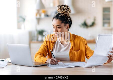 Arbeiten Sie von zu Hause aus. Konzentrierte, clevere afroamerikanische Frau, Studentin oder Designerin, in stilvoller Freizeitkleidung, in der Ferne studieren oder arbeiten, mit einem Laptop arbeiten, Blaupausen ansehen und untersuchen Stockfoto