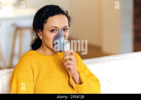 Medizinisches Behandlungskonzept. Kranke, erschöpfte Mischrassenfrau, Brünette, die im Wohnzimmer auf der Couch im gelben Pullover sitzt, behandelt wird, durch einen Inhalator atmet, die Kamera anschaut Stockfoto