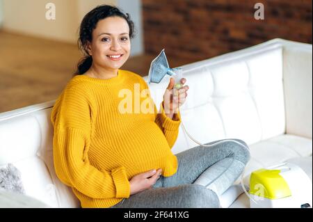 Freudig erholt sich schwanger gemischtes Rennen Brünette Frau, sitzt im Wohnzimmer auf der Couch tragen einen gelben Pullover, tut vorbeugende Behandlung, hält einen Inhalator in der Hand, Blick auf die Kamera Stockfoto