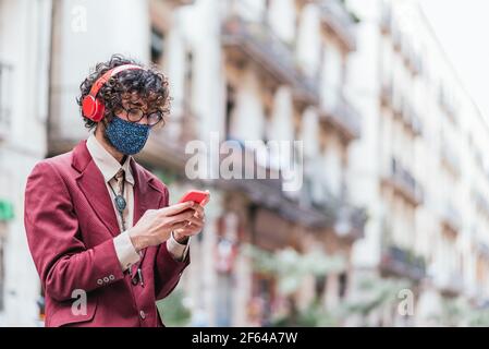 Horizontales Porträt eines kaukasischen schlanken jungen Mannes mit seinem Handy. Er kleidet sich mit einer roten Jacke und trägt rote Kopfhörer und eine gemusterte blaue Gesichtsmaske. In den Straßen von Barcelona Stockfoto