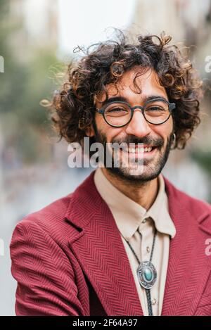 Nahaufnahme eines attraktiven kaukasischen jungen Mannes, der auf der Straße lächelt. Er hat lockiges Haar, Bart und Brille. Modische Freizeitkleidung Stockfoto