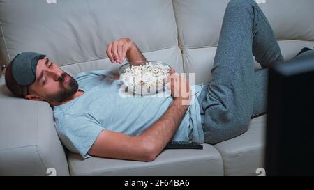 Müder Mann, der sich auf dem Sofa vor dem Fernseher entspannt und Popcorn isst, während er sich eine Filmvorstellung ansieht. Kaukasischer Mann mit Bart suchen Entertainment-Serie spät in der Nacht in der Küche Stockfoto