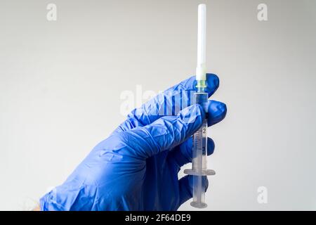 Die Hand des medizinischen Mitarbeiters in einem blauen Operationshandschuh hält die Spritze vor einem weißen Hintergrund. Nahaufnahme einer Chirurgen-Hand mit einem Impfstoff. Eine Spritze mit dem Stockfoto
