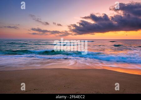 meeresstrand mit dramatischem Himmel bei Sonnenaufgang. Wunderschöne Urlaubslandschaft. Wellen Rollen auf dem Sand im Morgenlicht Stockfoto