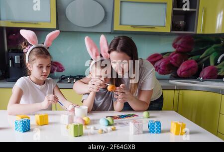 Glückliche Familie: Mutter Tochter und Sohn mit Hasenohren bereiten sich auf den Urlaub vor und färben Eier in der gemütlichen Küche des Hauses. Stockfoto