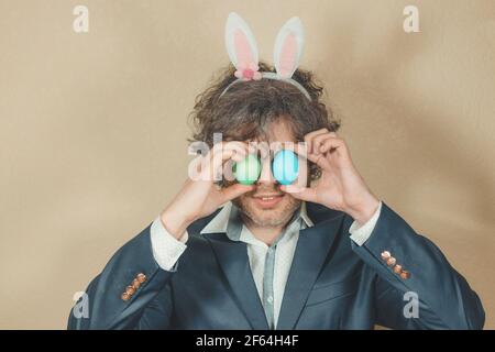 Ein lächelnder Mann in einem Anzug mit Hasenohren bedeckte seine Augen mit bunten Eiern. Fröhliches und fröhliches Osterkonzept. Weichfokus. Abgeschwächt Stockfoto