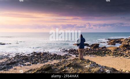 Ein Panoramablick auf einen Mann, der auf Felsen steht und auf das Meer bei Little Fistral in Newquay in Cornwall blickt. Stockfoto