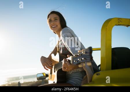 Fröhliche kaukasische Frau, die auf dem Strandbuggy am Meer sitzt Gitarre spielen Stockfoto