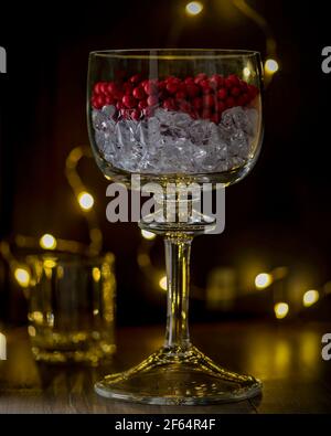 Eine elegante Glasvase ist mit einer Schicht von gefüllt Eiswürfel und eine Schicht roter Beeren gegen ein Dunkler Hintergrund mit Lichterketten Stockfoto