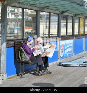 Boscombe, Bournemouth, Dorset, Großbritannien, 30.. März 2021, Wetter. Hitzewelle im Frühling. Ein älteres Paar sitzt beim Lesen der Zeitungen am Pier. Stockfoto