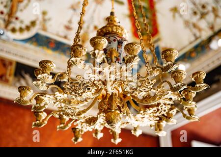 Nahaufnahme eines antiken goldenen italienischen Lüsters. Auf dem Hintergrund der bemalten Decke hängt ein Kronleuchter mit vergoldeten Ketten. Stockfoto