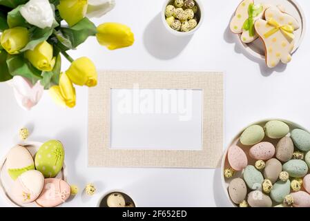 Ostertisch-Setting mit einem Teller aus farbigen Pastelleiern, einem Bouquet von Tulpen und Keksen Kaninchen, Fotorahmen für Mockup auf hellem Hintergrund Stockfoto