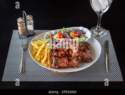 Glasierte Grillrippen mit Pommes Frites und einem gesunden Salat. Besteck und ein Glas Wasser, für eine Mahlzeit aufgestellt. Stockfoto