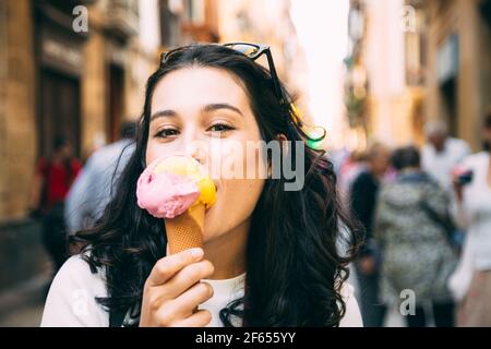 Glückliche junge schöne Frau genießt ein großes Eis auf der Straße. Nicht erkennbare Personen im Hintergrund Stockfoto