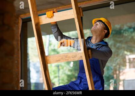 Junge männliche Baumeister in Overalls und Harthut suchen konzentriert, Klettern die Leiter während der Arbeit auf Hütte Baustelle Stockfoto