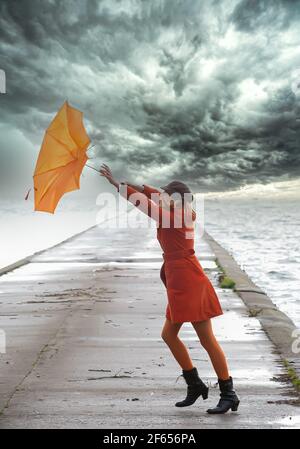 Ein Mädchen in orangefarbenem Mantel mit Regenschirm bei stürmischem Wetter auf dem Pier. Wolkiger Himmel. Stockfoto