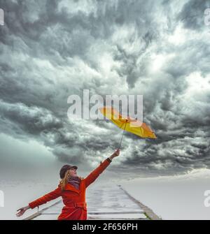 Ein Mädchen in orangefarbenem Mantel mit Regenschirm bei stürmischem Wetter auf dem Pier. Wolkiger Himmel. Stockfoto