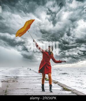 Ein Mädchen in orangefarbenem Mantel mit Regenschirm bei stürmischem Wetter auf dem Pier. Wolkiger Himmel. Stockfoto