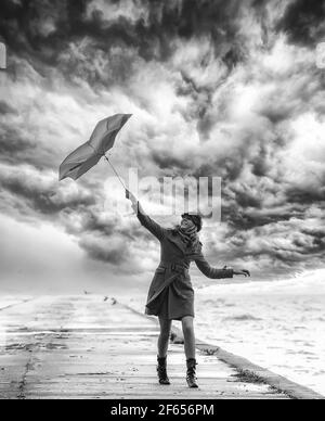 Ein Mädchen in orangefarbenem Mantel mit Regenschirm bei stürmischem Wetter auf dem Pier. Wolkiger Himmel. Schwarz und Weiß. Stockfoto
